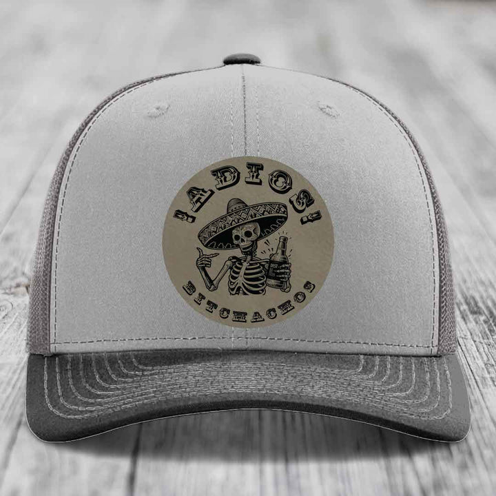 Grey trucker hat with circular leather patch featuring skeleton in sombrero holding beer bottle and text &