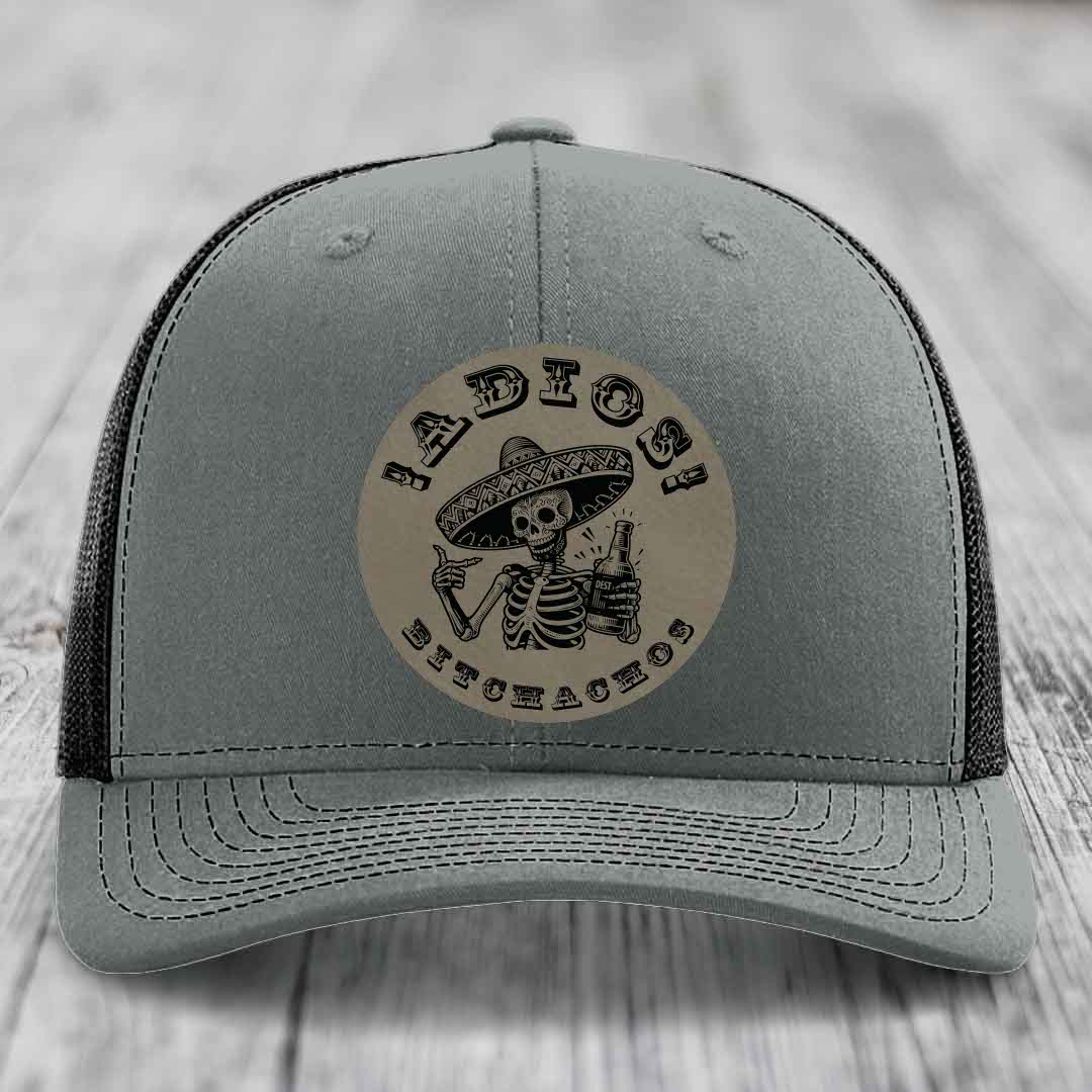 Grey trucker hat with black mesh back and a circular leather patch featuring a skeleton in a sombrero holding a beer bottle with &
