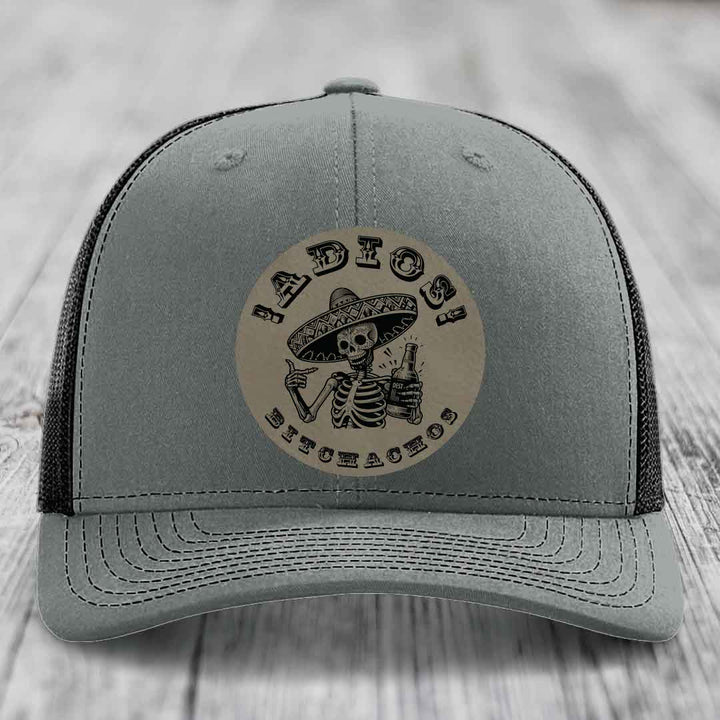 Grey trucker hat with black mesh back and a circular leather patch featuring a skeleton in a sombrero holding a beer bottle with &