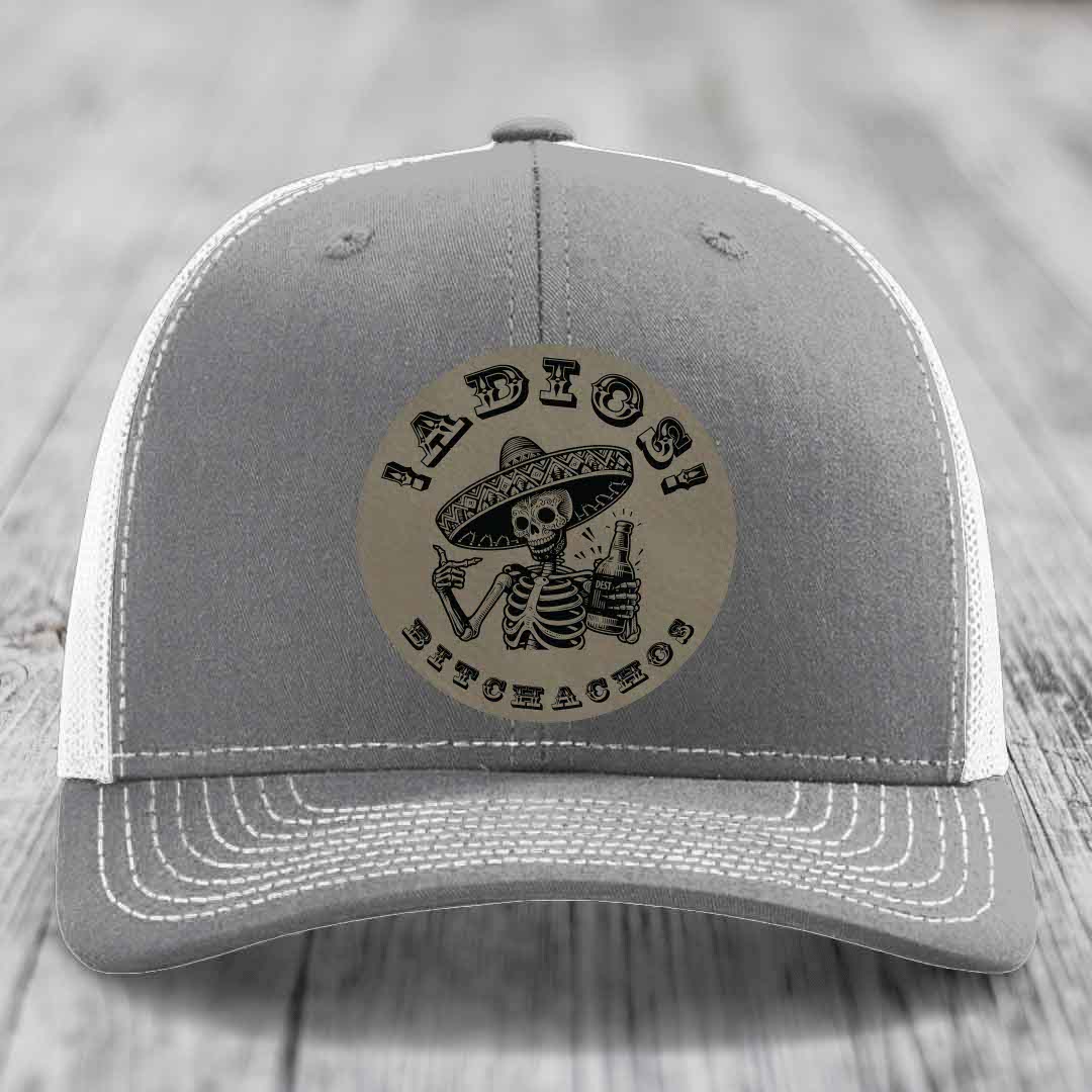 Grey trucker hat with white mesh back and stitching, featuring a circular leather patch of a skeleton in a sombrero holding a beer bottle with &