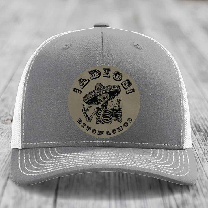 Grey trucker hat with white mesh back and stitching, featuring a circular leather patch of a skeleton in a sombrero holding a beer bottle with &