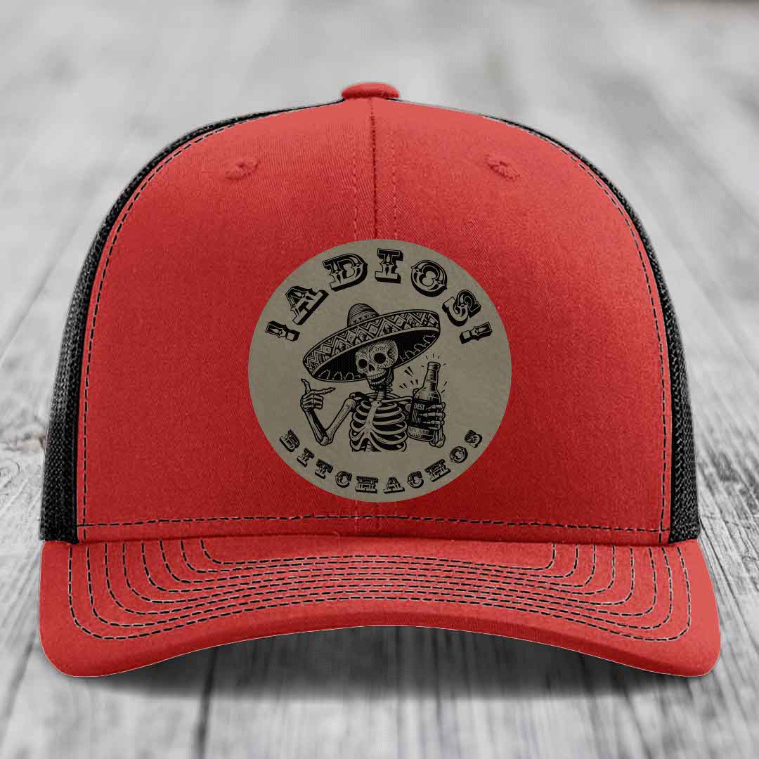 Red trucker hat with black mesh back and gray circular patch featuring a skeleton in a sombrero holding a beer bottle with text &