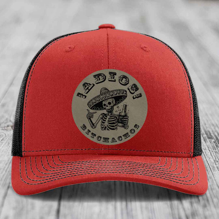 Red trucker hat with black mesh back and gray circular patch featuring a skeleton in a sombrero holding a beer bottle with text &