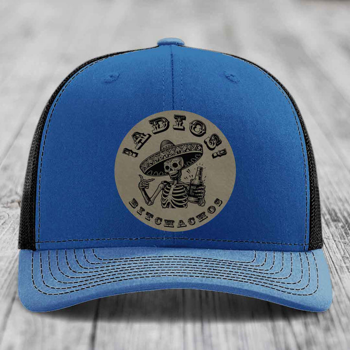 Blue trucker hat with black mesh back and beige circular patch featuring a skeleton in a sombrero holding a beer bottle.