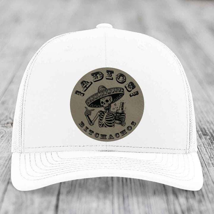 White trucker hat with a circular leather patch featuring a skeleton in a sombrero holding a beer bottle and the text &