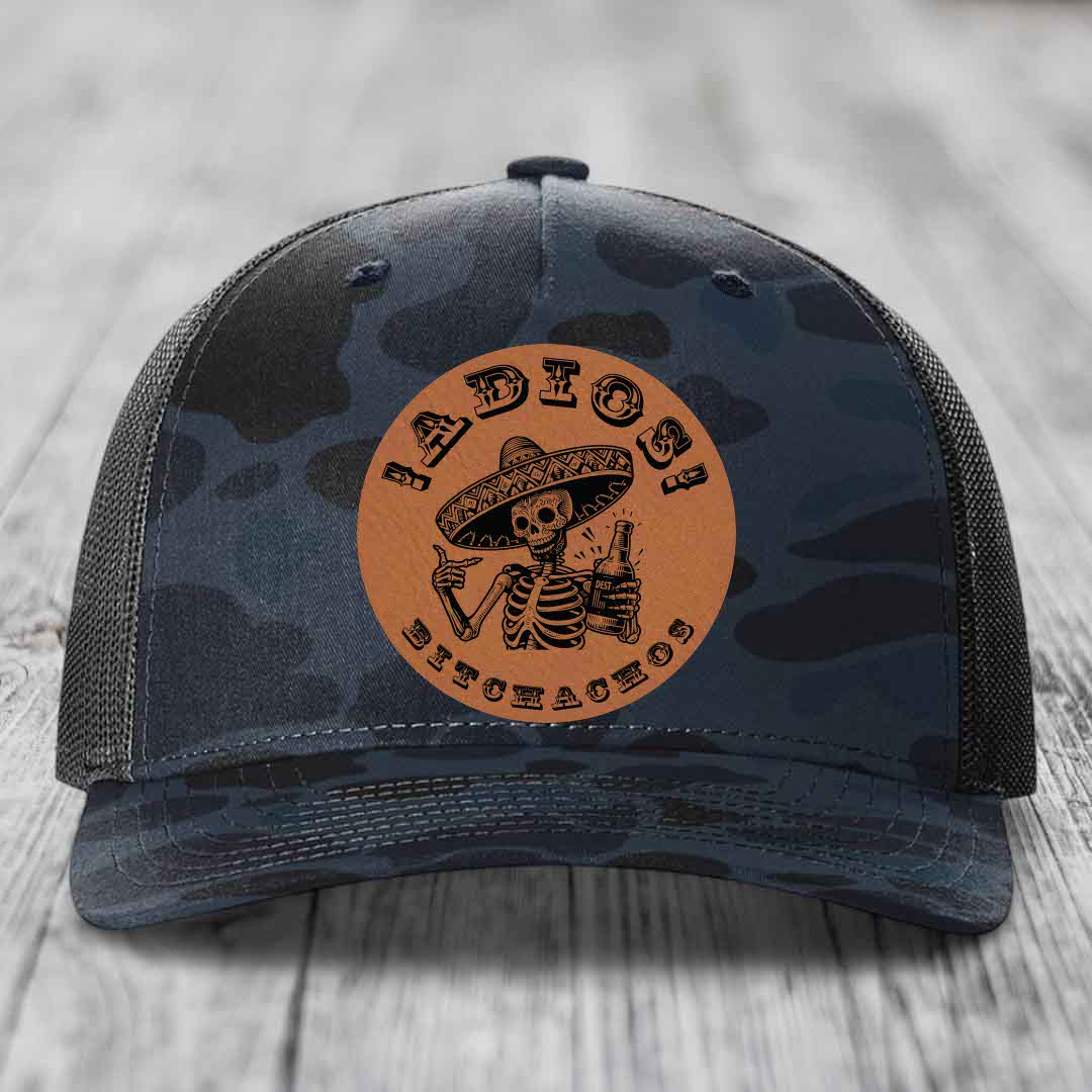 Camouflage trucker hat with brown leather patch featuring skeleton in sombrero holding beer bottle.