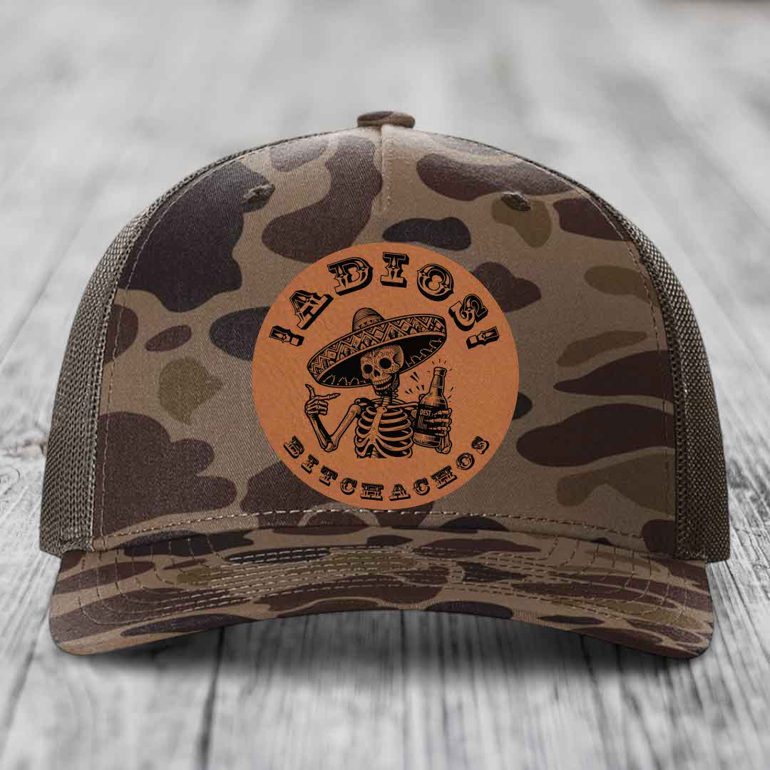 Camouflage trucker hat with brown leather patch featuring skeleton in sombrero holding beer bottle.