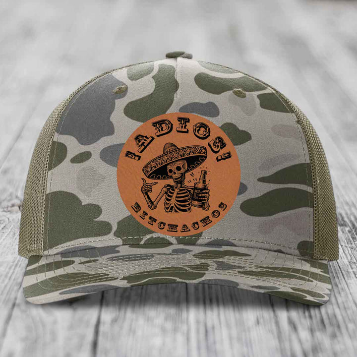 Camouflage trucker hat with brown leather patch featuring skeleton in sombrero holding beer bottle.