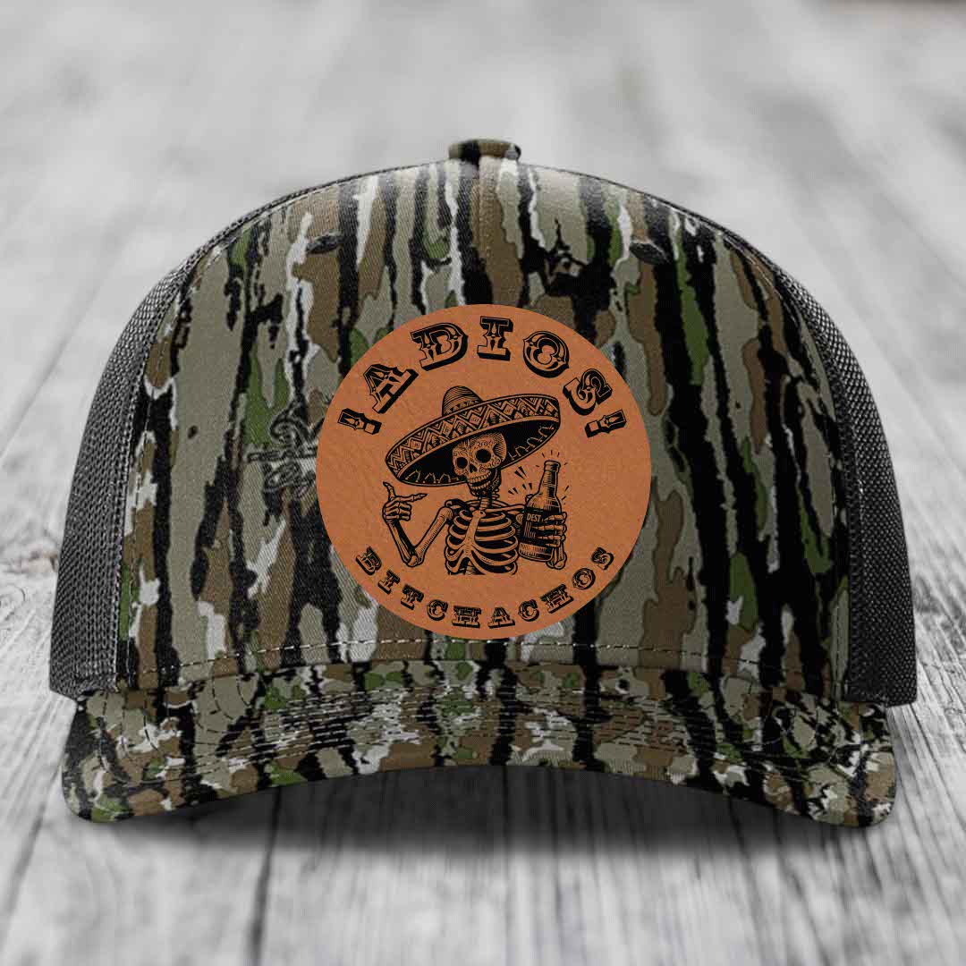 Camouflage trucker hat with brown leather patch featuring skeleton in sombrero holding beer bottle.