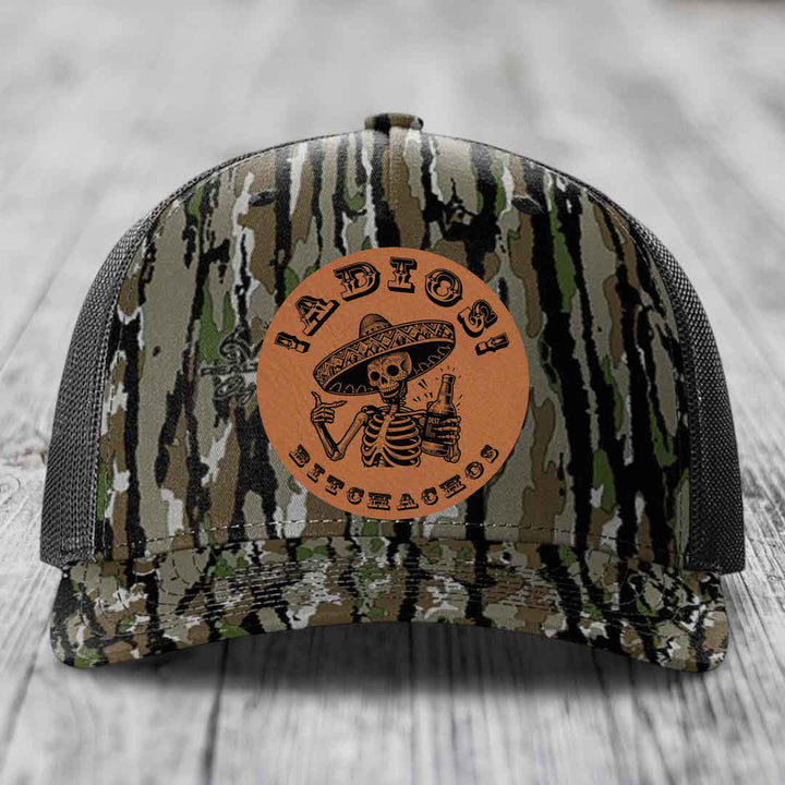 Camouflage trucker hat with brown leather patch featuring skeleton in sombrero holding beer bottle.