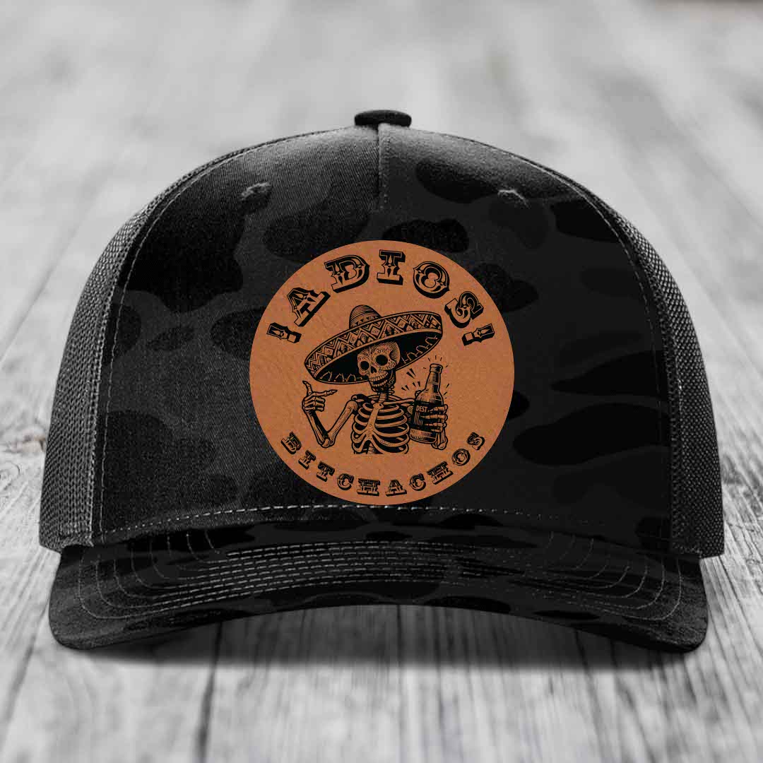 Black camouflage trucker hat with brown circular patch featuring skeleton in sombrero holding beer bottle.