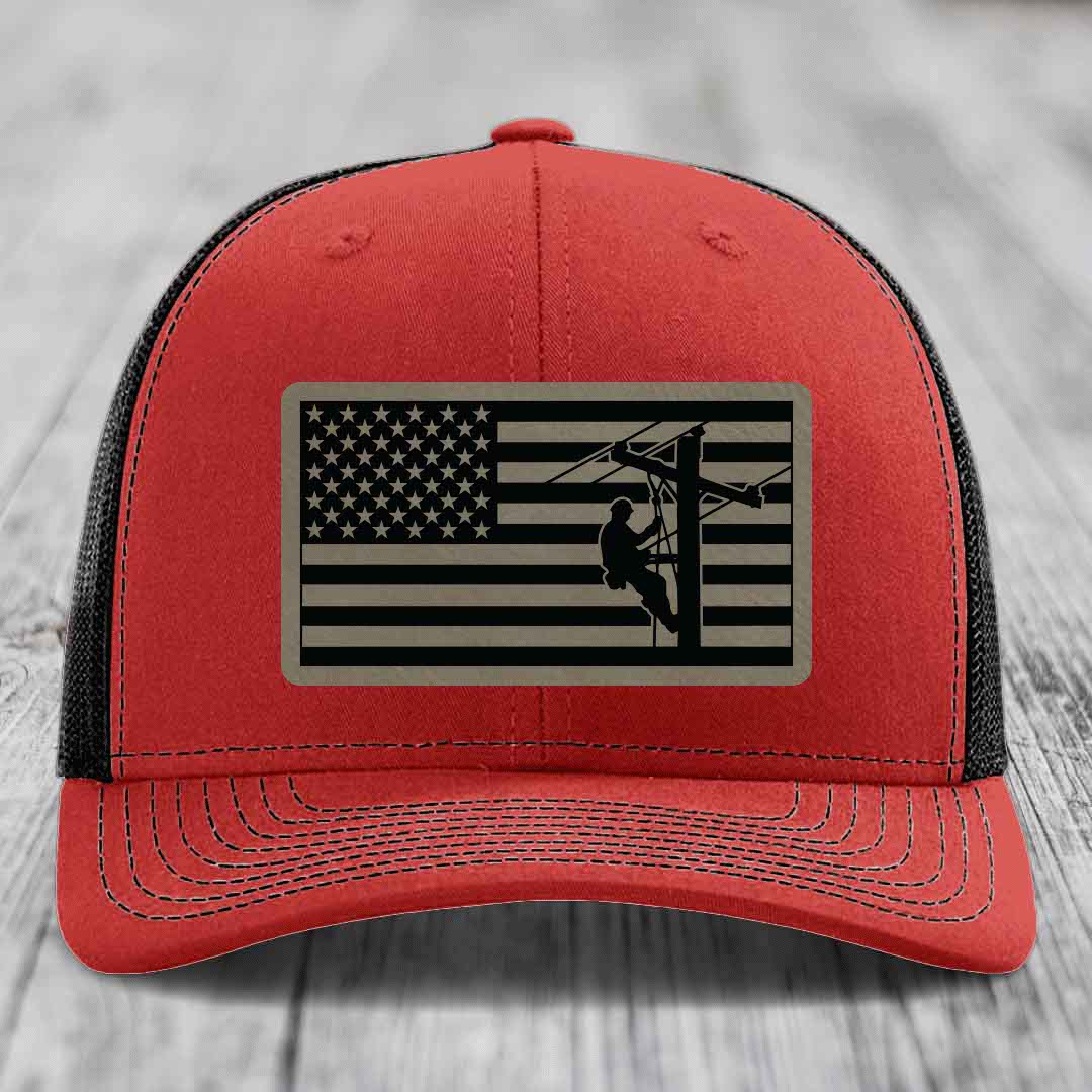 Red trucker hat with black mesh back and a rectangular American flag patch featuring a lineman silhouette.