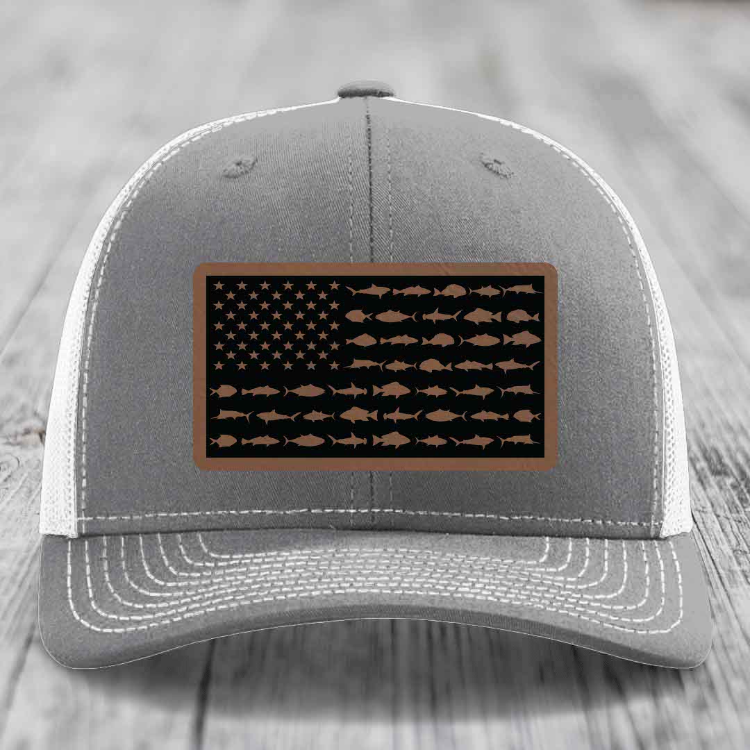 Grey trucker hat with white mesh back and stitched details, featuring a rectangular leather patch with an American flag design made of fish silhouettes.