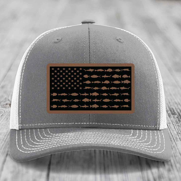 Grey trucker hat with white mesh back and stitched details, featuring a rectangular leather patch with an American flag design made of fish silhouettes.