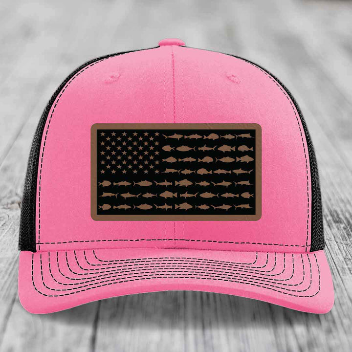 Pink trucker hat with black mesh back, featuring a rectangular leather patch with an American flag design made of fish silhouettes.