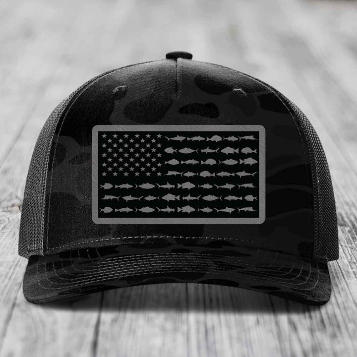 Black camouflage trucker hat with American flag patch made of saltwater fish silhouettes.