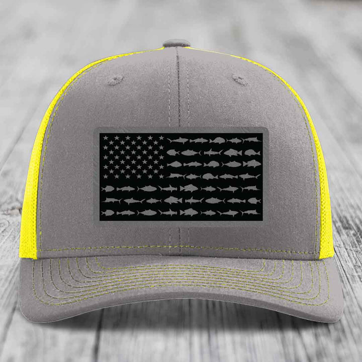 Grey trucker hat with yellow mesh and stitching, featuring a black American flag patch made of saltwater fish silhouettes.