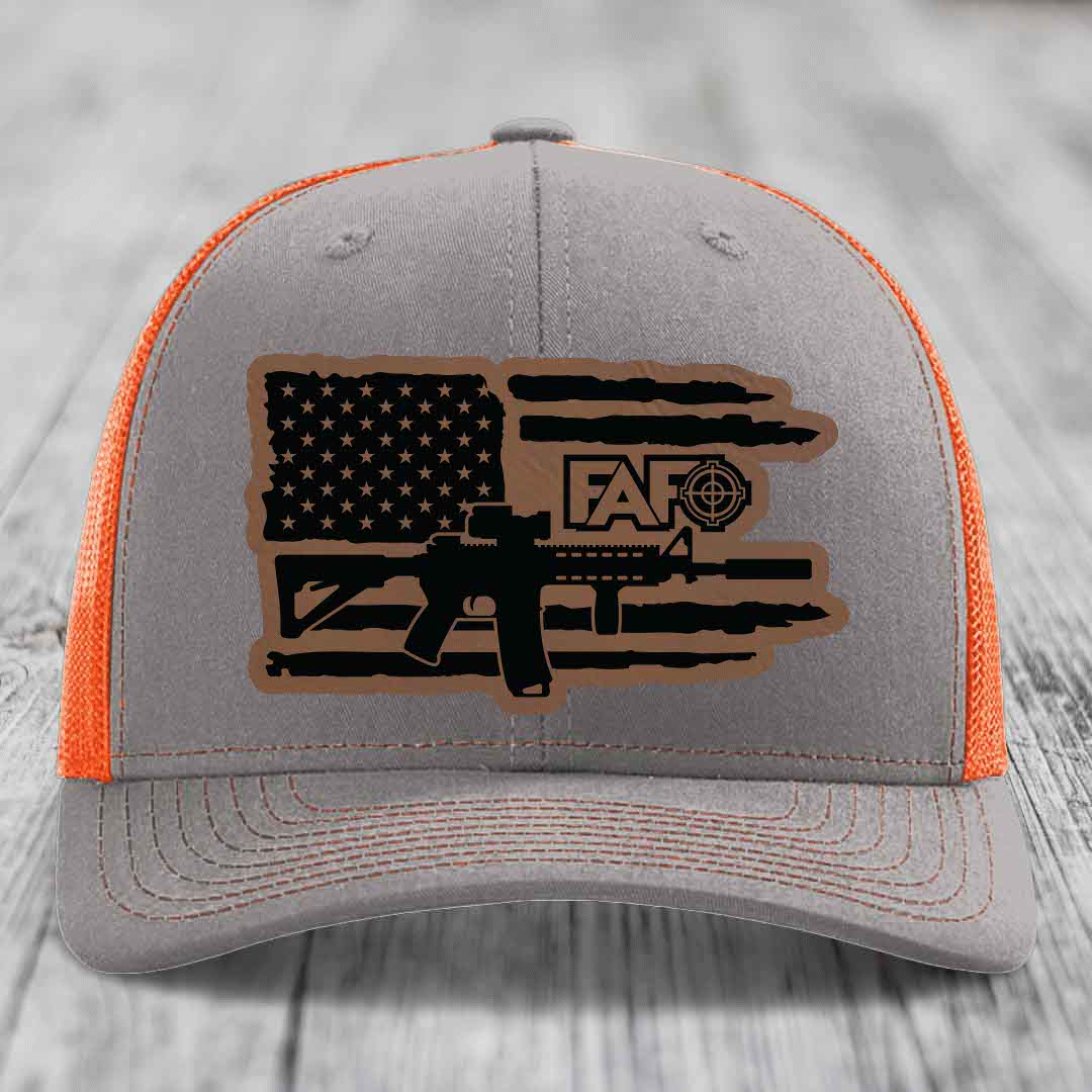 Grey trucker hat with orange mesh, featuring a brown leatherette patch of American flag, AR-15 rifle, and &