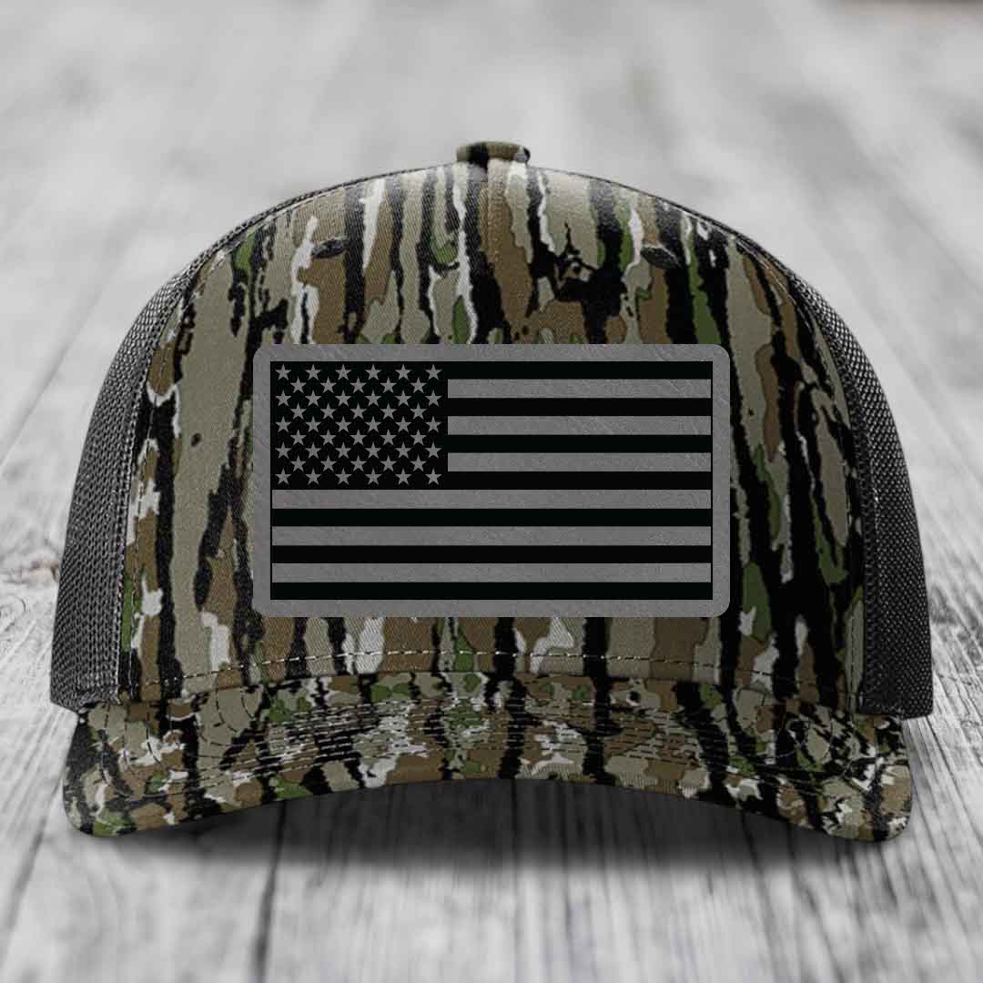 Camouflage trucker hat with American flag patch on front, black mesh back, resting on wooden surface.