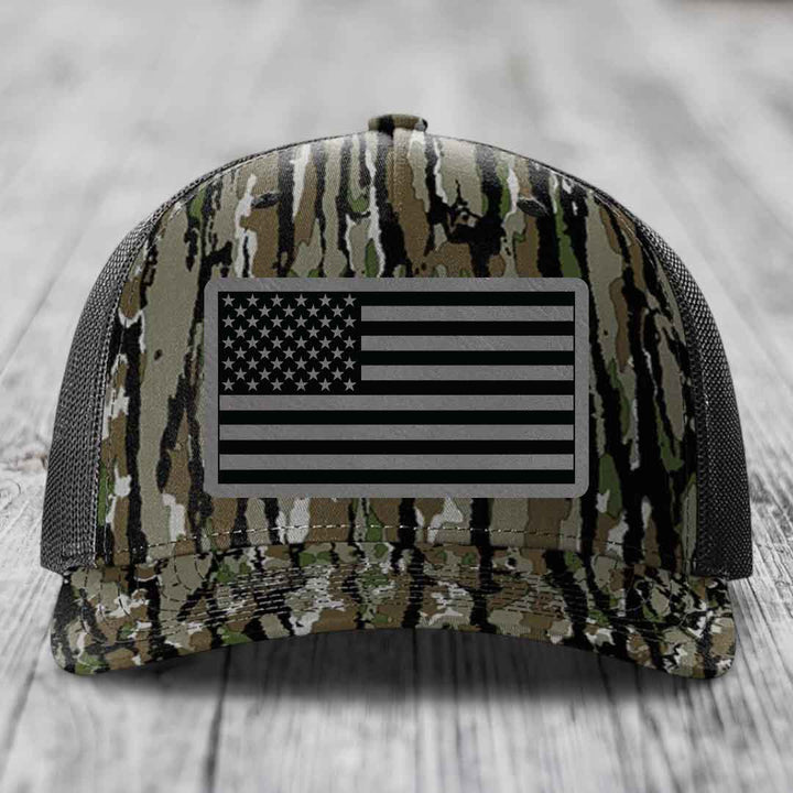 Camouflage trucker hat with American flag patch on front, black mesh back, resting on wooden surface.