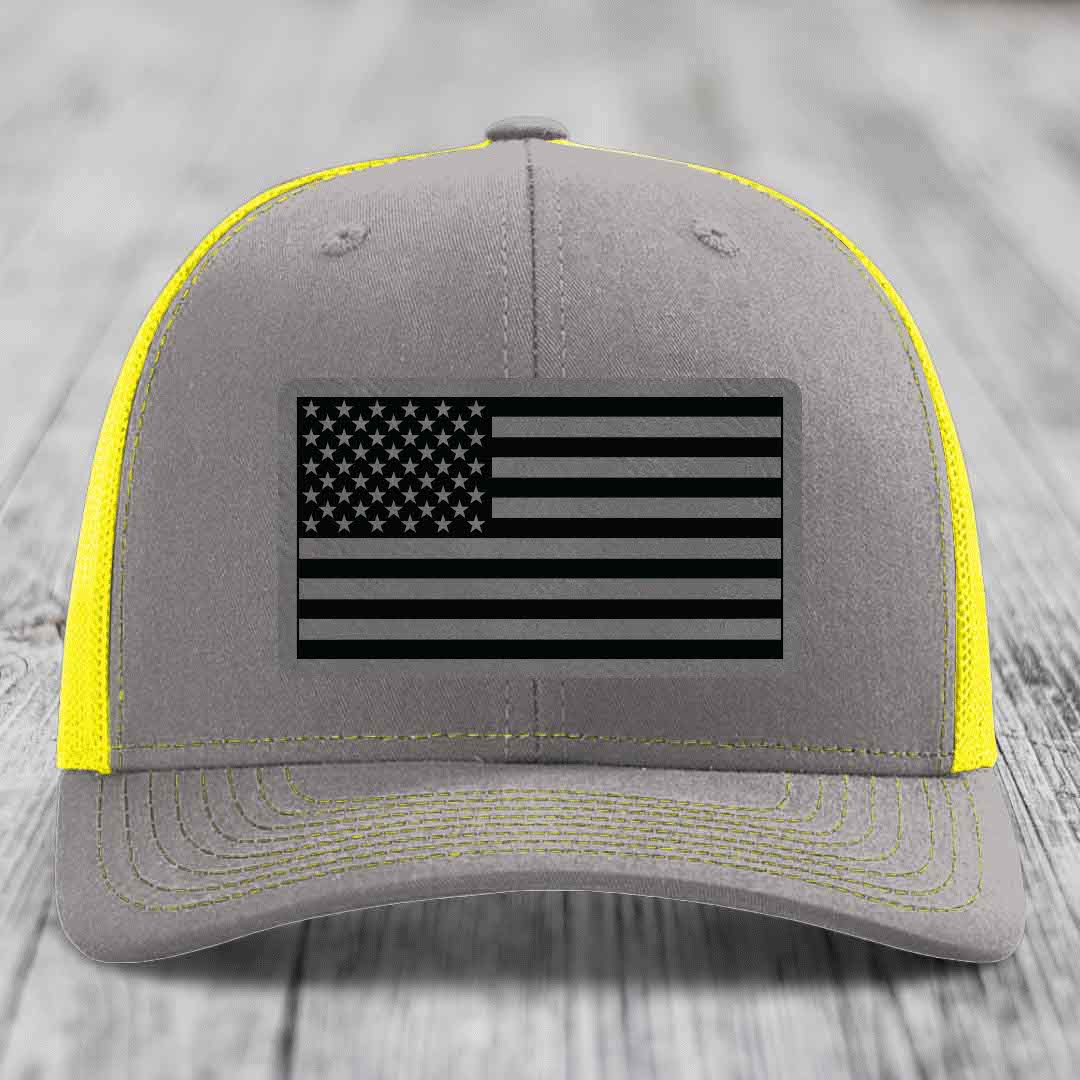 Grey trucker hat with yellow mesh and stitching, featuring a black and grey American flag patch.