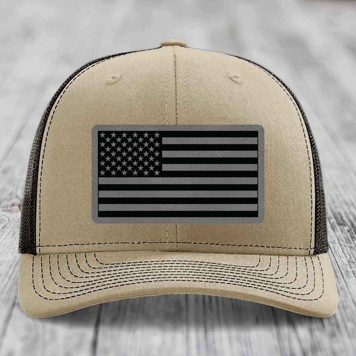 Tan trucker hat with black and grey American flag patch on front.