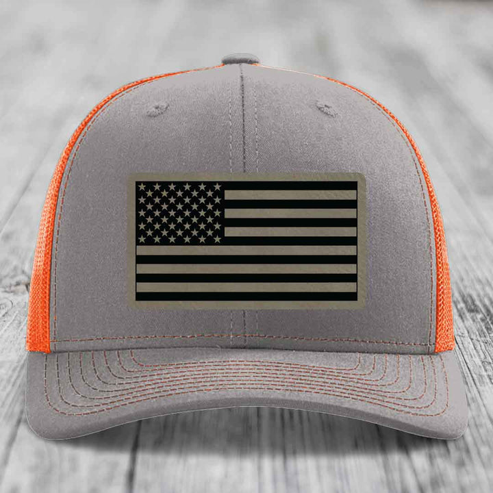 Grey trucker hat with orange mesh back and black American flag patch on front.