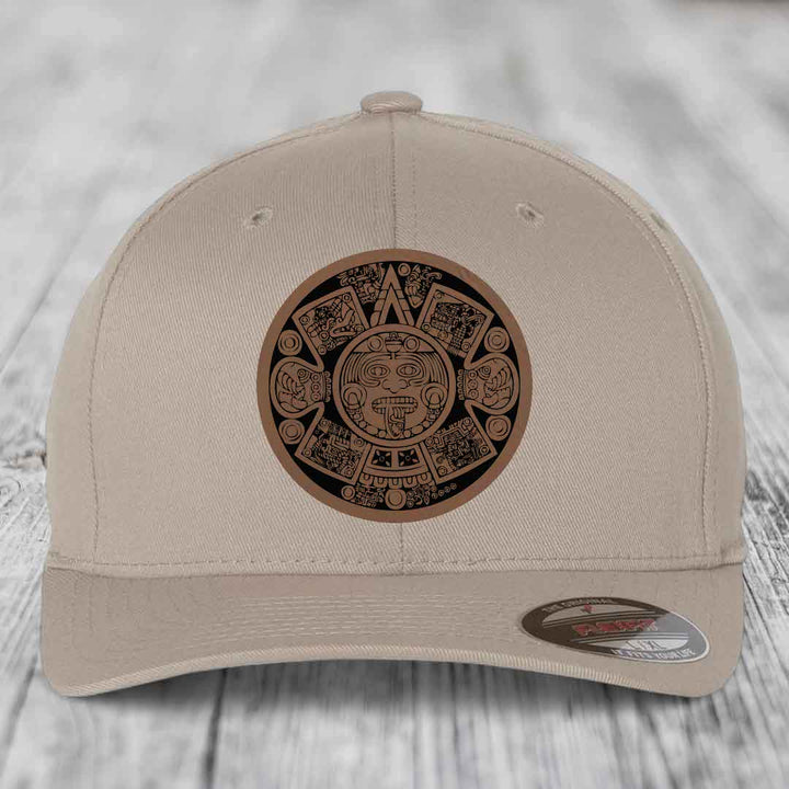 Beige baseball cap with Aztec calendar leather patch on front, displayed on wooden surface.