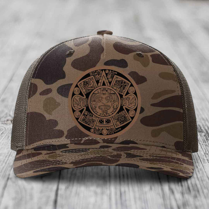 Camouflage trucker hat with Aztec calendar patch on front, placed on wooden surface.
