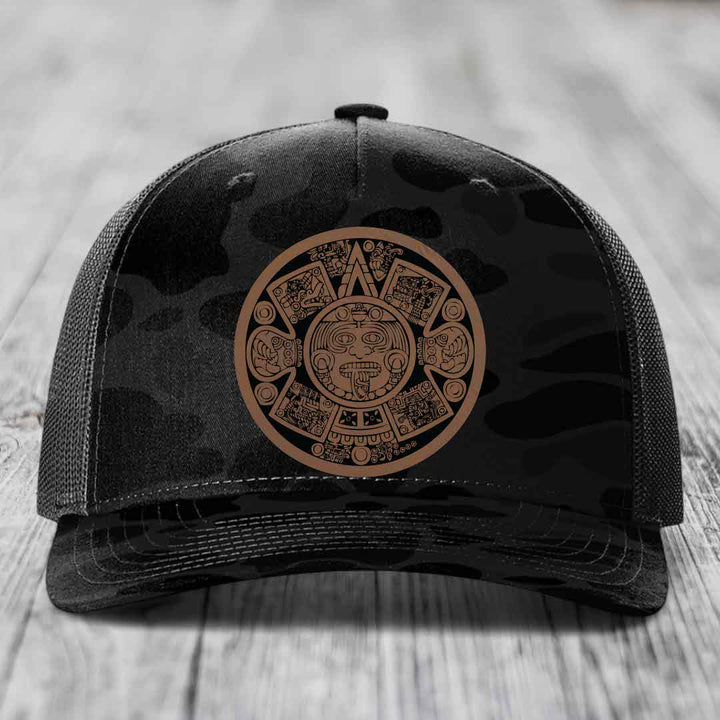 Black trucker hat with brown Aztec calendar patch on front, placed on wooden surface.