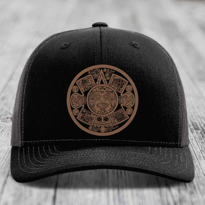 Black trucker hat with brown Aztec calendar patch on front, placed on wooden surface.