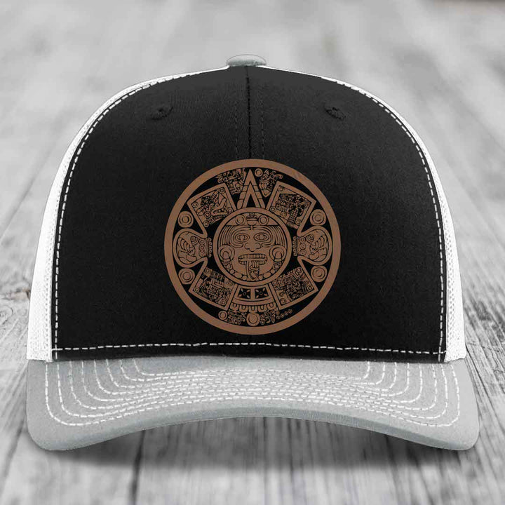 Black and grey trucker hat with brown Aztec calendar patch on front.