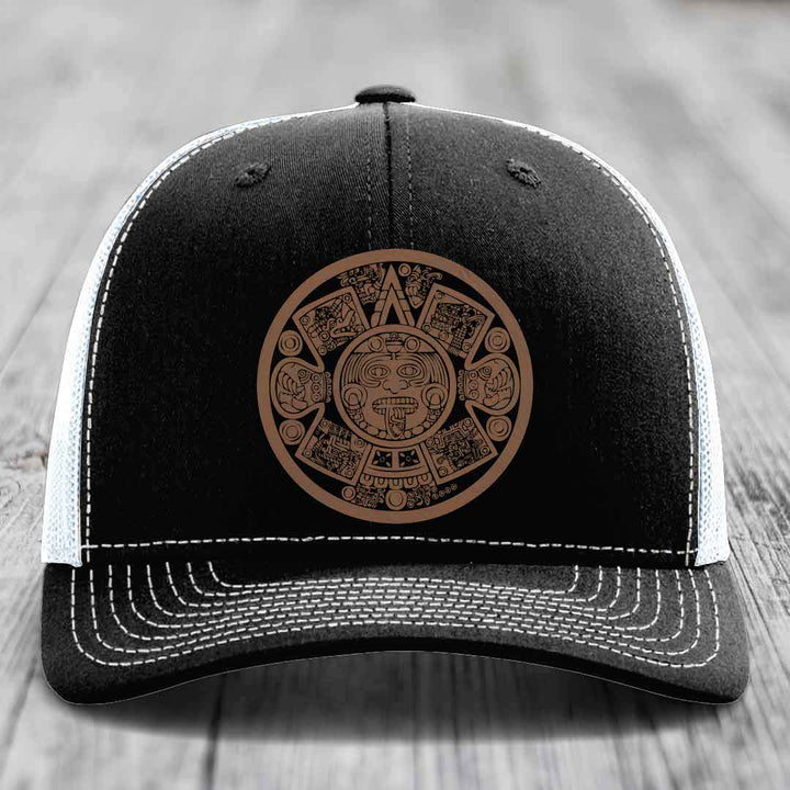 Black trucker hat with brown Aztec calendar patch and white stitching on front panel.