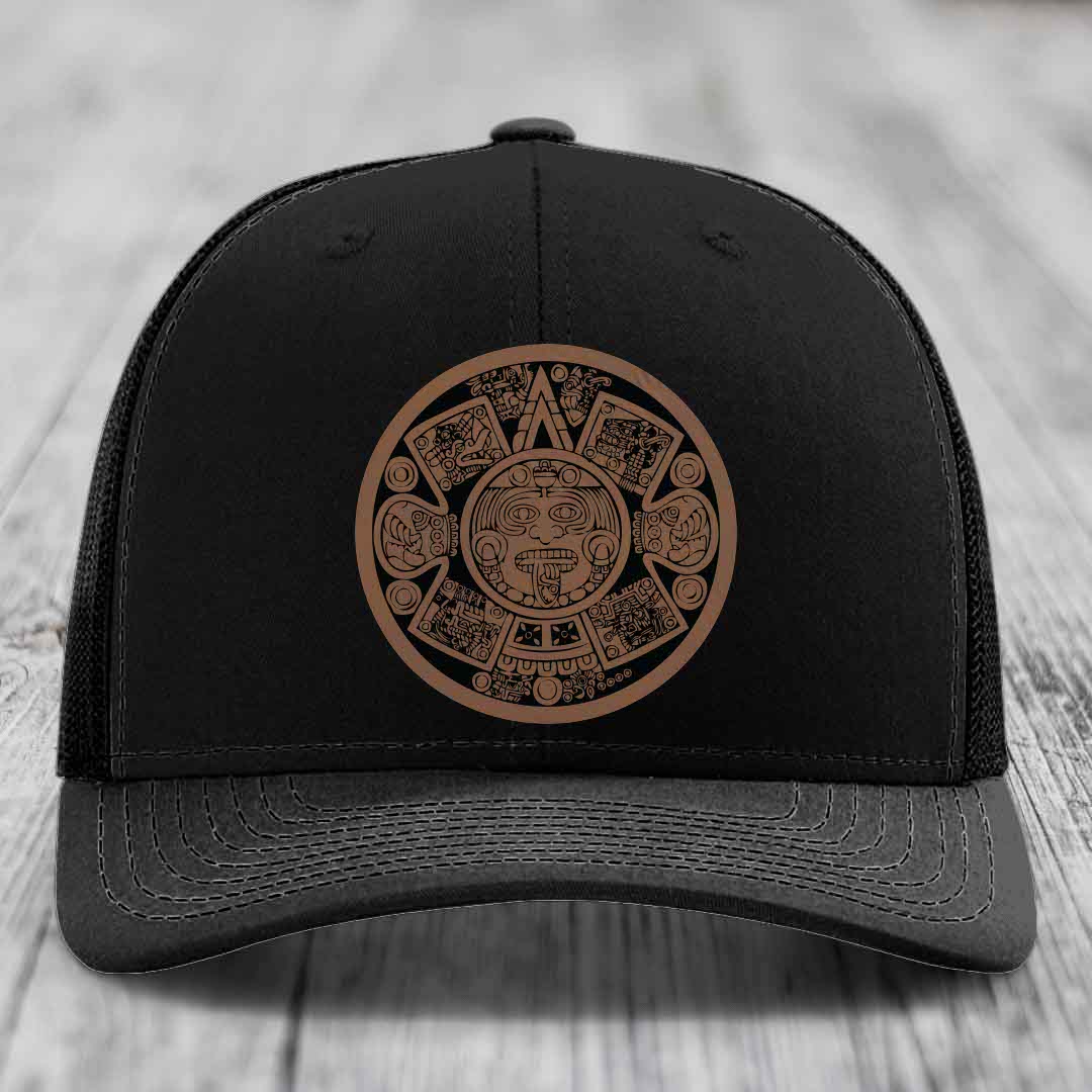 Black trucker hat with brown Aztec calendar patch on front, placed on wooden surface.