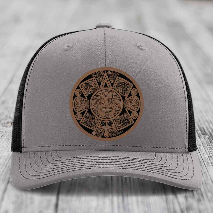Grey trucker hat with brown Aztec calendar leather patch on front.