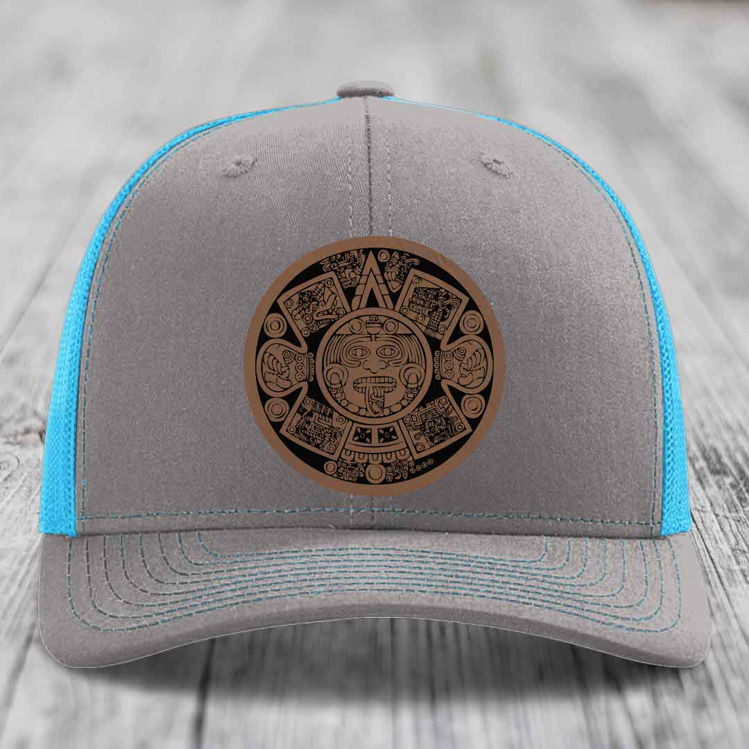 Grey trucker hat with blue mesh sides and a brown Aztec calendar leather patch on front.