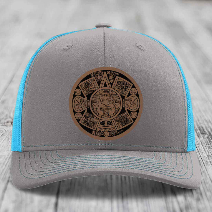 Grey trucker hat with blue mesh sides and a brown Aztec calendar leather patch on front.