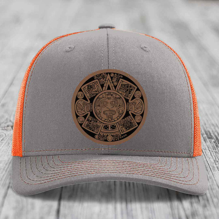 Grey trucker hat with orange mesh back and brown Aztec calendar leather patch on front.
