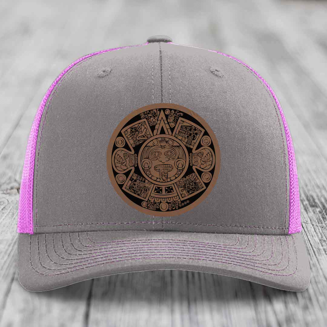 Grey trucker hat with pink mesh and a brown Aztec calendar leather patch on front.