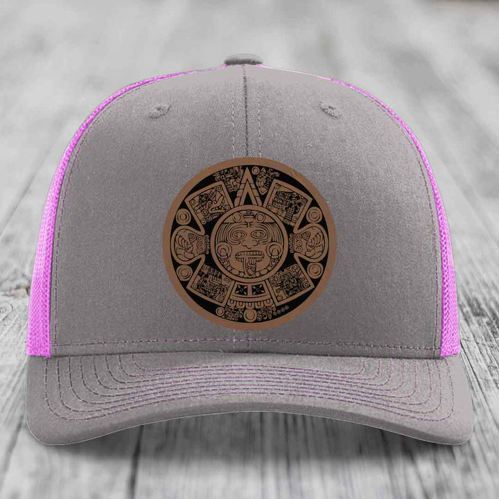 Grey trucker hat with pink mesh and a brown Aztec calendar leather patch on front.