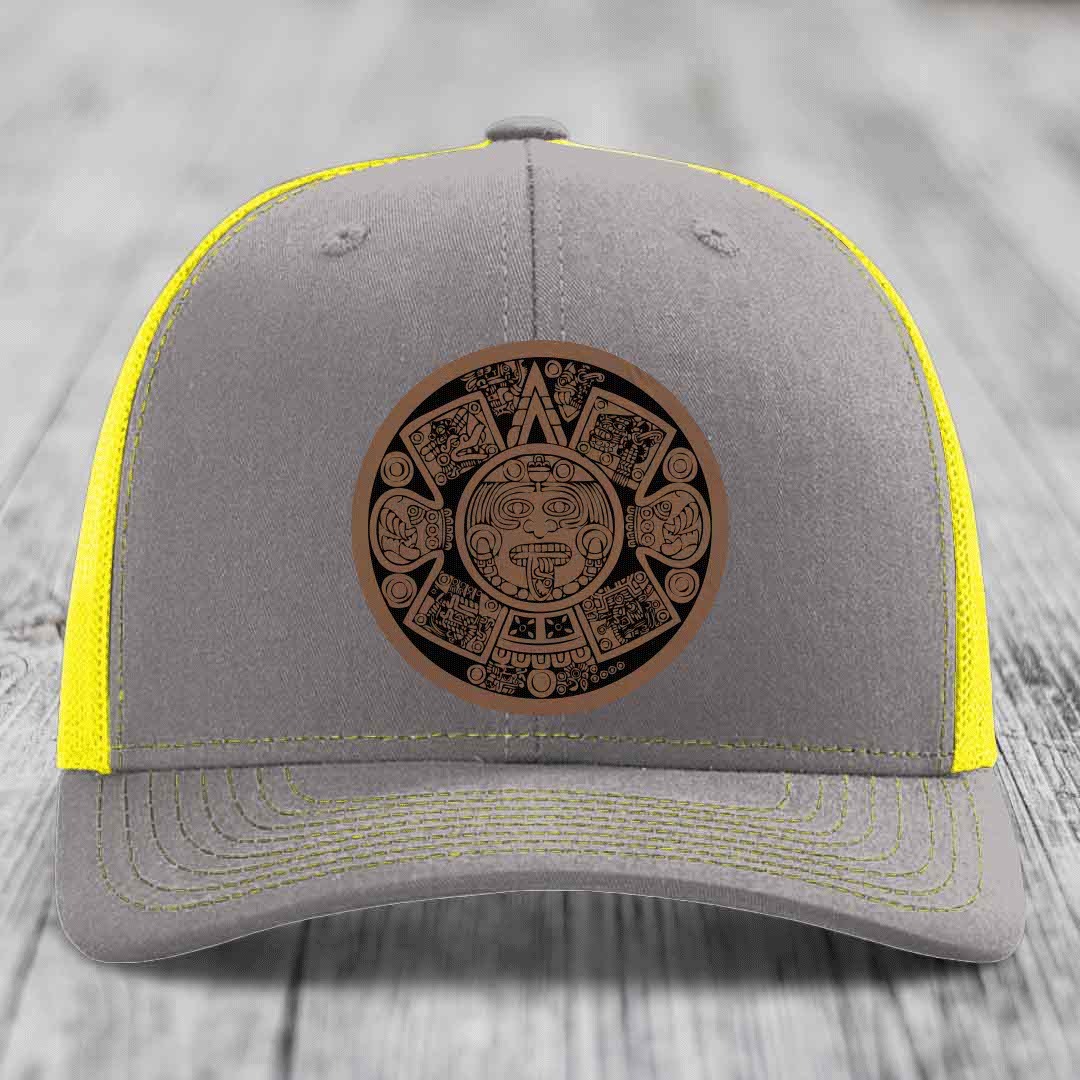 Grey trucker hat with yellow mesh and stitching, featuring a brown Aztec calendar leather patch.