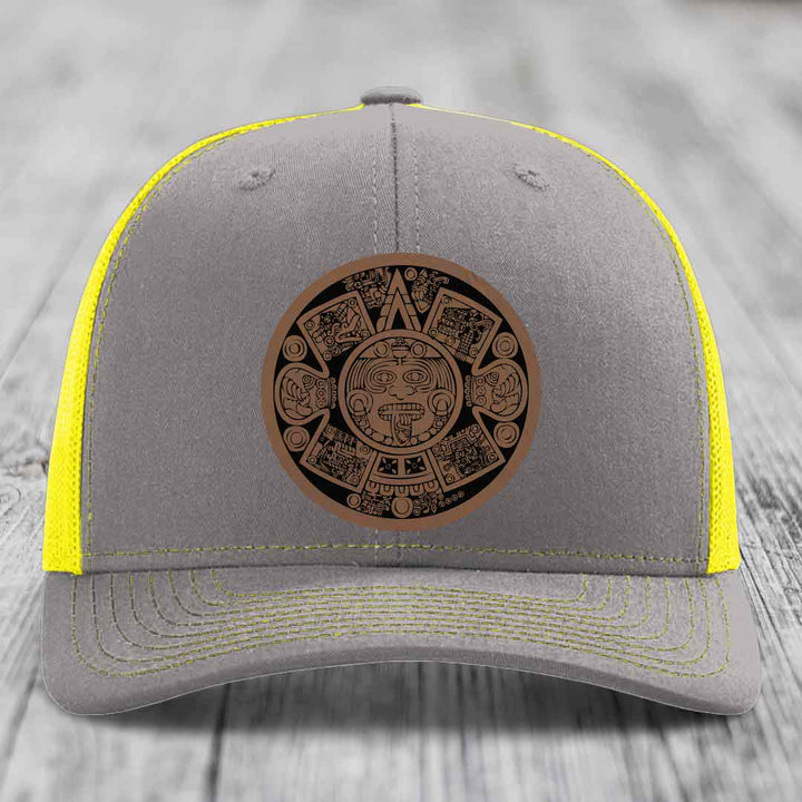 Grey trucker hat with yellow mesh and stitching, featuring a brown Aztec calendar leather patch.