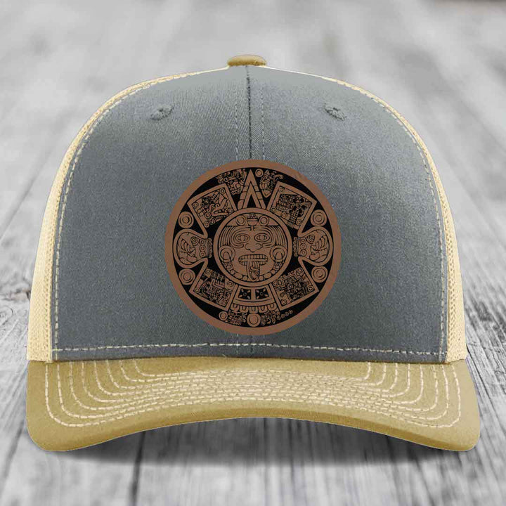 Grey and beige trucker hat with Aztec calendar leather patch on front.