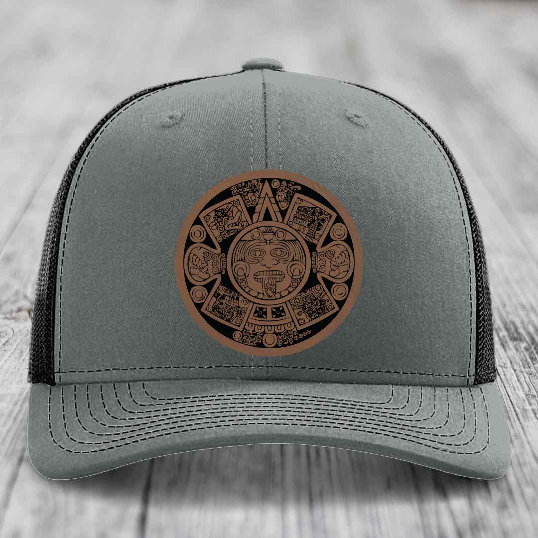 Grey trucker hat with brown Aztec calendar leather patch on front.
