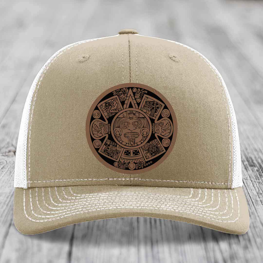 Tan trucker hat with white mesh back and embroidered Aztec calendar patch on front.