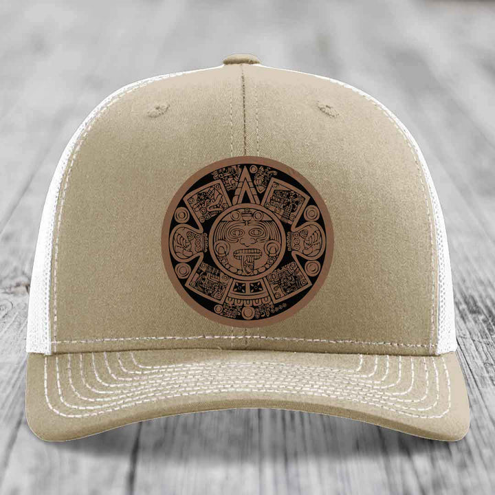 Tan trucker hat with white mesh back and embroidered Aztec calendar patch on front.