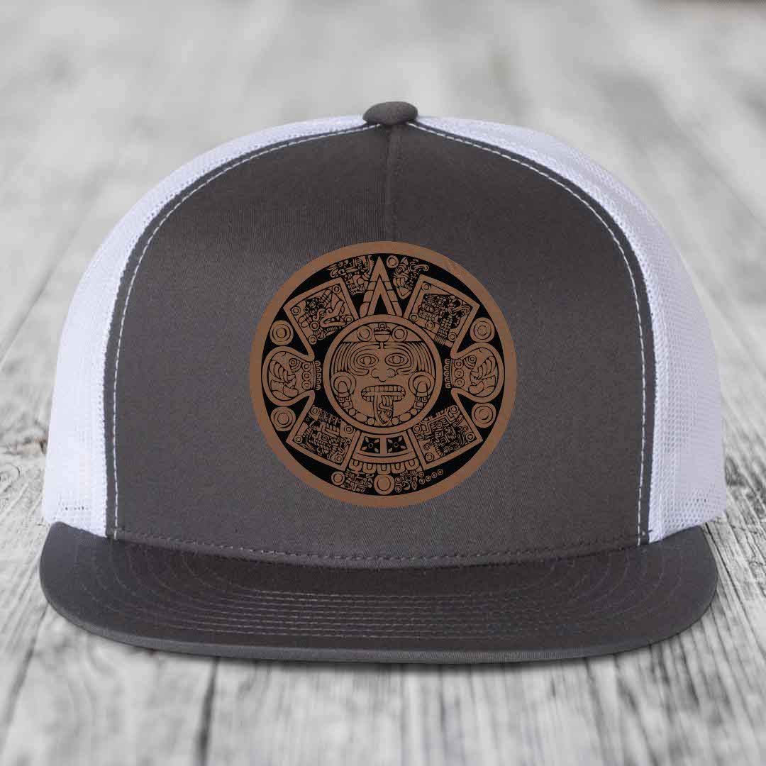 Grey front panel trucker hat with brown Aztec calendar patch, white mesh back, and grey brim.
