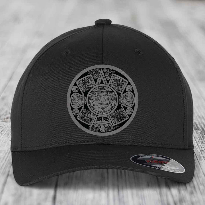 Black baseball cap with Aztec calendar design on front, displayed on wooden surface.