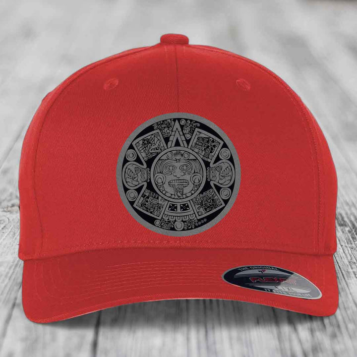Red baseball cap with Aztec calendar patch on front, displayed on wooden surface.