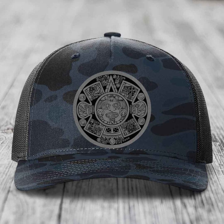 Camouflage trucker hat with Aztec calendar patch on front, placed on wooden surface.